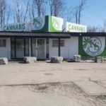 Small cannabis shop with green signage in a rural area, surrounded by bare trees and a large empty parking lot.