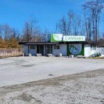 Small cannabis shop with green signage in a rural area, surrounded by bare trees and a large empty parking lot.