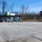 Small cannabis shop with green signage in a rural area, surrounded by bare trees and a large empty parking lot.
