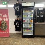 Bright cannabis retail interior with glass display cases, product shelves, and logo decals on the windows and KS merch hanging on a rack and a banner of Pink Kush