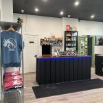 Bright cannabis retail interior with glass display cases, product shelves, and logo decals on the windows and KS merch hanging on a rack