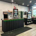 Bright cannabis retail interior with glass display cases, product shelves, and logo decals on the windows.