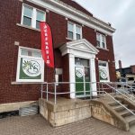 Red-brick cannabis store with green 'KS' logos, a red 'CANNABIS' flag, and a green door marked 'OPEN' at the center entrance.