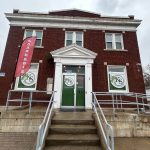 Red-brick cannabis store with green 'KS' logos, a red 'CANNABIS' flag, and a green door marked 'OPEN' at the center entrance.