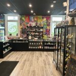 Bright cannabis retail interior with glass display cases, product shelves, and logo decals on the windows.
