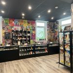 Bright cannabis retail interior with glass display cases, product shelves, and logo decals on the windows.