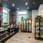 Bright cannabis retail interior with glass display cases, product shelves, and logo decals on the windows.